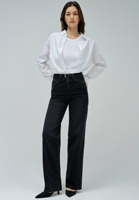 Mujer vistiendo una camisa blanca de botonadura sobre una camiseta blanca, con unos pantalones vaqueros de tiro alto y pernera ancha negros, cinturón negro y tacones puntiagudos negros.