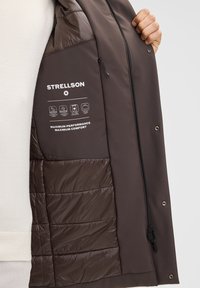 Person, der eine offene braune Strellson-Jacke hält und das gepolsterte Futter sowie die Etiketten für wasserdichte, winddichte und leichte Reiseeigenschaften zeigt.