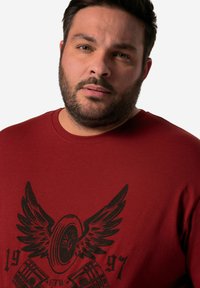 Červené bavlněné tričko s černým grafickým designem křídel a pneumatiky, se textem "1997" a "FTW" ve spodní části. Kulatý výstřih.