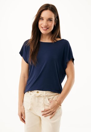 Lächelnde Frau mit langen braunen Haaren, die ein marineblaues kurzärmeliges Oberteil und beige Hosen mit geflochtenem Gürtel trägt, steht vor einem weißen Hintergrund.