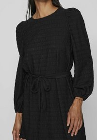 Una modelo lleva un vestido negro texturizado con cintura ceñida y mangas abullonadas, destacando los detalles y el ajuste de la prenda.