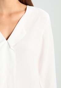 Blouse blanche avec un col en V et un col drapé subtil. Tissu lisse et léger avec un léger éclat. Manches longues et coupe décontractée.