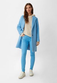 Cappotto di lana azzurro chiaro sopra una felpa beige, abbinato a pantaloni blu coordinati e sneakers bianche. Presenta un design sartoriale e dettagli con bottoni.