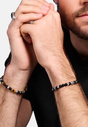 Man wearing black beaded bracelets with silver accents on both wrists and rings on fingers, hands clasped near face with partial beard visible.