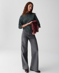 Vrouw met golvend bruin haar in een donkergroene, gestructureerde top en grijze, wijde jeans, die een opgevouwen bordeauxrode tas tegen een lichte achtergrond houdt.