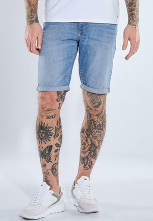 Beine mit verschiedenen schwarzen Tätowierungen, die hellblaue hochgekrempelte Jeansshorts, weiße Sneaker und ein weißes Hemd tragen.