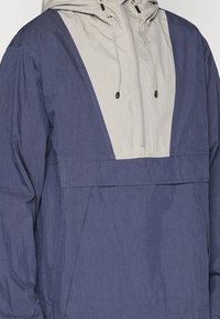 Navy blue anorak jacket with a light gray panel, featuring a front pocket, adjustable hood, and cinch drawstrings. Wrinkled texture.