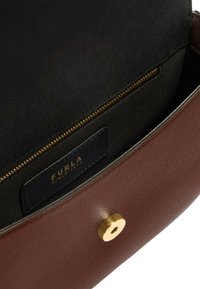 Interno di borsa in pelle marrone con tasca con zip, dettagli in oro e fodera in tessuto nero. Presenta un'etichetta con la scritta "Furla Since 1927."