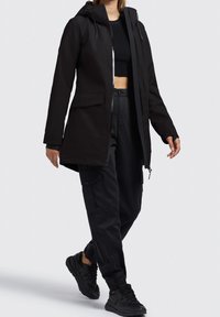 Sort vandtæt jakke med lynlås foran, justerbar hætte og to forlommer. Kombineret med sorte cargo bukser og sporty sorte sko.