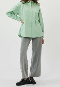 Chemise boutonnée vert clair avec une poche, coupe ample ; associée à un pantalon large gris clair et des chaussures à plateforme noires. Fond uni.