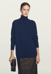 Donna che indossa un maglione dolcevita blu navy e una gonna marrone scuro, tenendo in mano sinistra una piccola borsa nera con texture.
