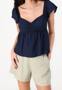 Navy blue short-sleeve blouse with a scoop neck, gathered detail at the bust, and a relaxed fit, paired with light beige shorts.