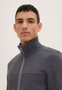 Siva zip-up jakna s visokim ovratnikom, koja ima glatku teksturu i kontrastni patentni zatvarač. Istaknuti šavovi duž područja prsnog koša.
