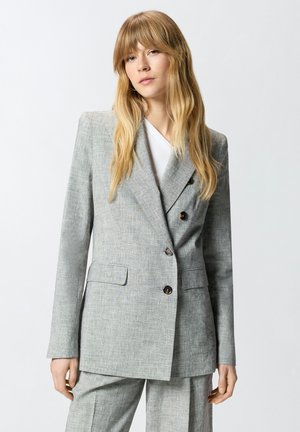 Donna bionda che indossa un completo doppiopetto grigio chiaro con bottoni neri e pantaloni abbinati, in piedi davanti a uno sfondo neutro.