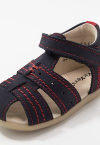 Sandalia de cuero azul marino con costuras rojas, diseño de punta abierta, correa ajustable y acentos recortados para ventilación. Forro interior suave.