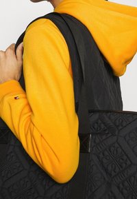 Schwarze, gesteppte Tragetasche mit stabilen Webbing-Griffen, die eine texturierte Oberfläche aufweist. Kombiniert mit einem leuchtend gelben Hoodie.