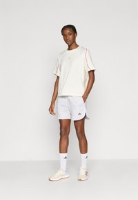 Kremna majica s kratkimi rokavi z rdečimi in zlatimi poudarki, združena s sivimi kratkimi hlačami. Bele nogavice in bež športni copati z oranžnimi podplati.