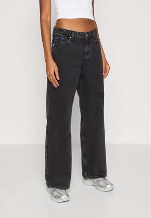 Person trägt locker sitzende, hochtaillierte schwarze Jeans und silberne Sneakers, kombiniert mit einem weißen bauchfreien, ärmellosen Top, steht auf einem grauen Boden.