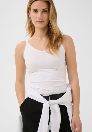 Jeune femme aux longs cheveux blonds portant un débardeur blanc et un pantalon noir avec une chemise blanche nouée autour de la taille.