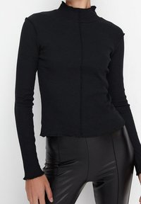Schwarzes, geripptes Langarmoberteil mit Stehkragen und rohem Saum. Der Stoff ist weich und hat eine figurbetonte Silhouette. Getragen mit schwarzen, glänzenden Hosen.