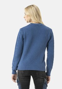 Pull en tricot bleu avec un motif texturé, des poignets et un ourlet côtelés, et des détails en tricot torsadé le long des manches. Porté sur un pantalon cargo noir.