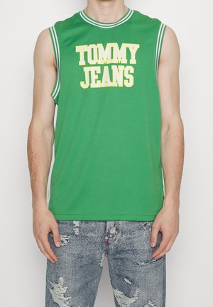 Mężczyzna w zielonym, sportowym topie bez rękawów z napisem "TOMMY JEANS" oraz zniszczonych niebieskich dżinsach, stojący z ramionami swobodnie opuszczonymi wzdłuż ciała.