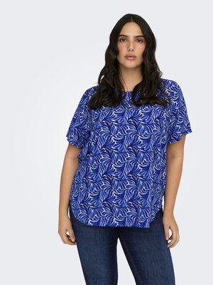 Vrouw met lang donker haar, die een blauw-wit gebloesd kort mouwen topje en donkerblauwe spijkerbroek draagt, staand tegen een effen achtergrond.