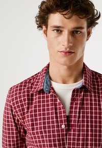 Camicia a quadri rossi e bianchi con colletto in denim. Presenta una chiusura con bottoni nella parte anteriore e una maglietta bianca sotto. Il tessuto sembra morbido e strutturato.