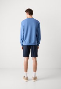 Light blue sweatshirt with long sleeves, paired with dark navy shorts. Model's shoes are beige, and he wears white ankle socks.