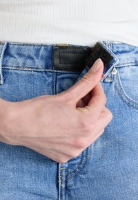 Blue denim jeans with a black waistband patch. A hand is adjusting a hook-and-loop fastener at the front. The fabric shows a faded texture.