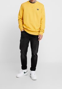 Žlutá mikina s kulatým výstřihem a černou logovou nášivkou, spojená s černými džíny a bílými teniskami s zelenými akcenty.