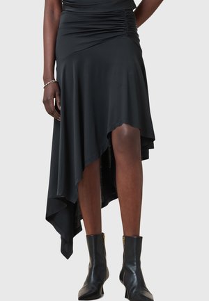 Jupe asymétrique noire en tissu extensible, dotée de détails froncés et d'une texture lisse. Portée avec des bottes noires hauteur cheville.