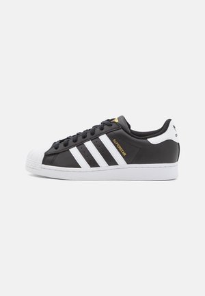 Czarny but sportowy Adidas Superstar z białymi paskami, wzmocnionym noskiem i białą podeszwą, pokazany z profilu na jasnym tle.