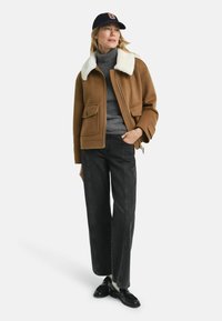 Frau, die eine braune Jacke mit weißen Kragen, einen grauen Rollkragenpullover, schwarze weite Hosen, schwarze Slipper und eine marineblaue Mütze trägt, steht mit einer Hand in der Tasche.