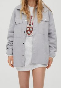 Grå ruskindsjakke med en afslappet pasform, trykknapper, to brystlommer og opkrøllede ærmer, båret over en grå sweatshirt.