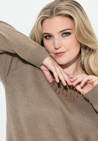 Brązowy sweter robiony na drutach z teksturowanym wzorem, ozdobiony pomarańczowymi i złotymi koralikami. Modelka pokazuje ręce w pobliżu dekoltu.