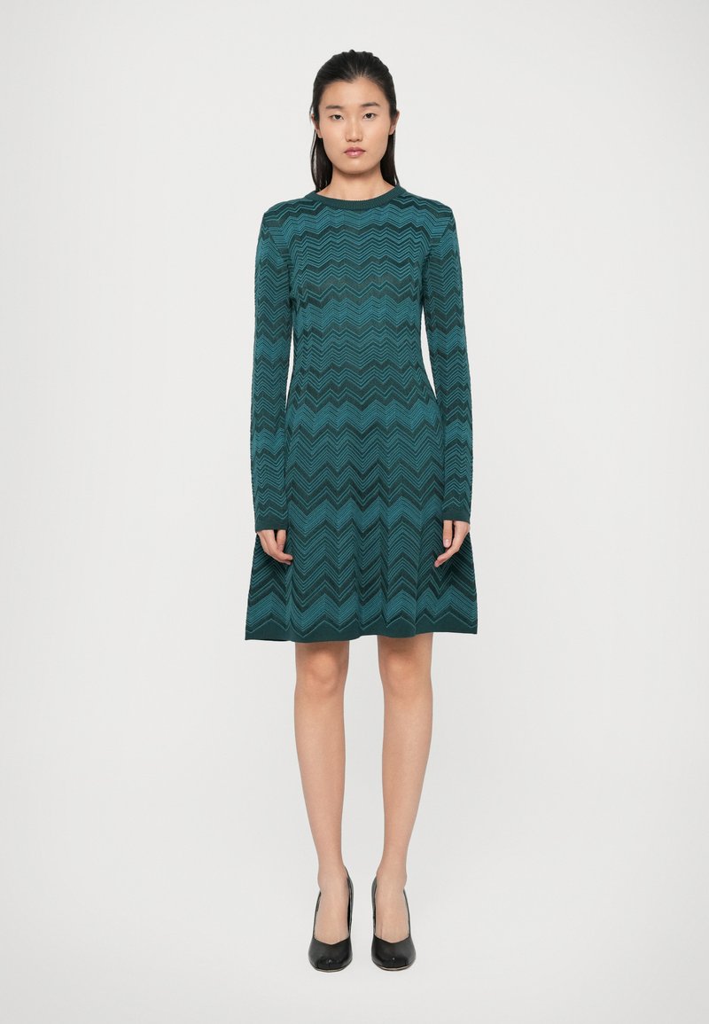 Robe longue teal à manches longues avec un motif en zigzag, corsage ajusté et jupe évasée, fabriquée en tissu tricoté, assortie à des chaussures à talons hauts noires.