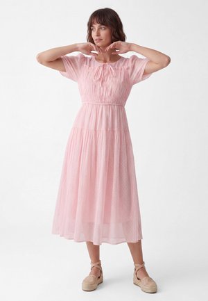 Kvinde i en lys pink, kortærmet, midi kjole med bindingsnakke og espadrillos poser med armene løftet nær ansigtet.