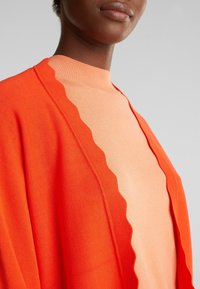 Femme portant un top pêche clair à col haut et un cardigan orange vif avec des bords festonnés sur un fond gris clair uni.