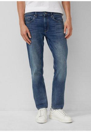 Person iført medium blå jeans med lige pasform og hvide sneakers står mod en neutral baggrund.