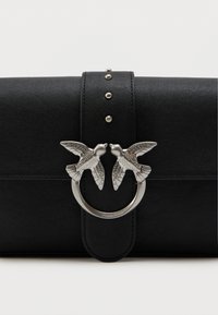 Czarna skórzana kopertówka z pionowym paskiem, zdobiona srebrnymi ćwiekami i okrągłym srebrnym zapięciem ozdobionym dwoma szczegółowymi ptakami zwracającymi się ku sobie.