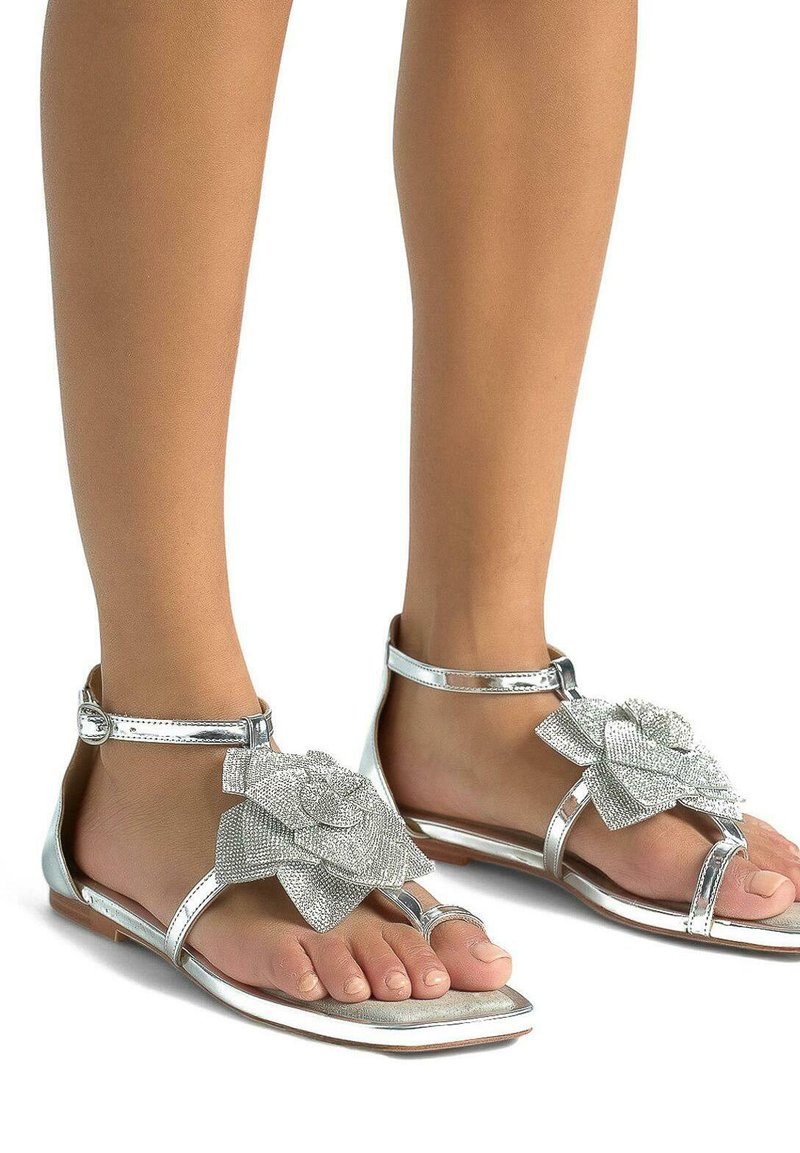 Sandali piatti argento con cinturini metallici e un grande fiore testurizzato come accento sulla punta. Finitura liscia, design open toe e fibbia alla caviglia.