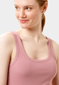 Světle růžový žebrovaný top bez rukávů se širokými ramínky, s kulatým výstřihem a hladkým povrchem. Detailní pohled na rameno modelky a výstřih.