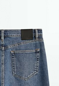 Bakfickan och midjan på blå denimjeans med synliga bältesöglor och en svart rektangulär lädermärkning.