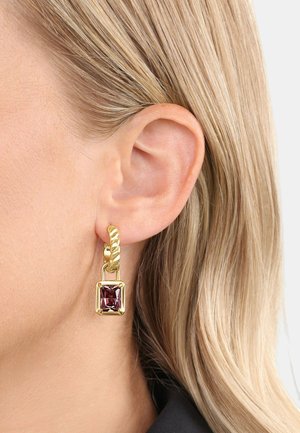 Boucle d'oreille créole torsadée en or avec une pierre précieuse carrée violette pendante portée sur une oreille blonde sur un fond uni.