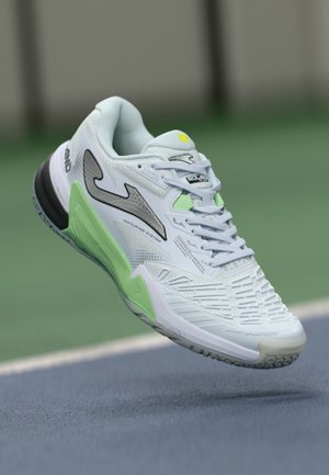 Chaussure de sport blanche et verte en plein air au-dessus d'une surface de court de tennis bleue, montrant les logos de la marque et les détails du système de ventilation.