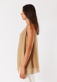 Top senza maniche, ampio, di colore marrone chiaro, realizzato in tessuto morbido e trasparente con un design drappeggiato e orlo arrotondato. Cuciture e dettagli minimi.