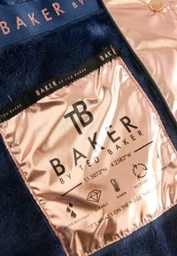 Navy-Fleece-Material zeichnet sich durch ein bronzefarbenes Metallschild mit Text und Symbolen aus, die Wasserbeständigkeit, Haltbarkeit, Wärme und recycelte Füllung anzeigen.