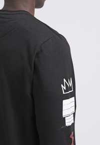 Maglietta nera a maniche lunghe con stampa di una corona bianca e un design astratto sulla manica sinistra; realizzata in morbido materiale di cotone.