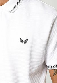 Polo shirt blanc avec texture côtelée, présentant un logo noir brodé et des accents à rayures noires contrastantes sur le col et les manches.